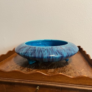May include: A low-profile, round ceramic bowl featuring a vibrant turquoise and deep blue drip glaze finish. The bowl is supported by three small, integrated feet and has a wide, open top with a smooth, glossy surface texture.