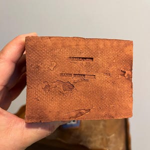 May include: A close-up of a textured, reddish-brown, rectangular block. The words "ARTOCA" and "HAND WORK" are stamped into the surface. The block is held in a person's hand.