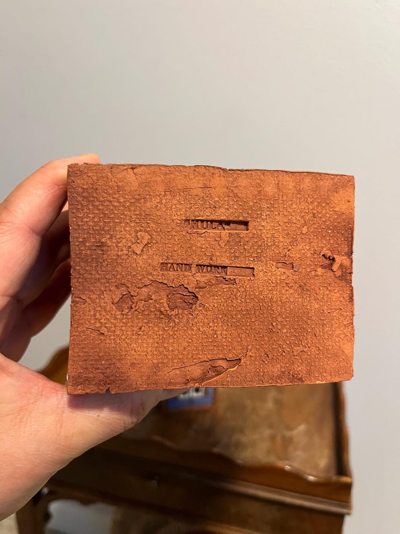 May include: A close-up of a textured, reddish-brown, rectangular block. The words "ARTOCA" and "HAND WORK" are stamped into the surface. The block is held in a person's hand.
