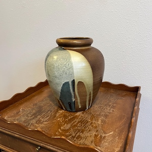 May include: A ceramic vase with a brown, beige, and blue-green color scheme. The vase has a rounded shape with a wide opening and a dripped glaze effect on one side. The vase is sitting on a wooden table.