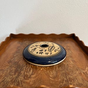May include: A ceramic, round, blue and tan decorative piece with a central hole. The blue rim features tan symbols. The piece is resting on a wooden tray with a scalloped edge. The background is a plain white wall.