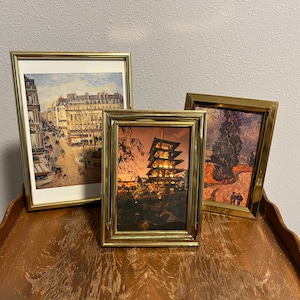 May include: Three rectangular picture frames with polished gold-tone metal borders. The left frame displays a Parisian street scene, the center frame shows a multi-tiered pagoda at dusk, and the right frame features a landscape with a tall cypress tree.