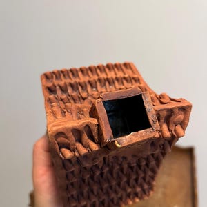 May include: A terracotta-colored ceramic house-shaped container with a textured roof and a square chimney. The container has a dark interior and is held in a person's hand. The house has decorative details.