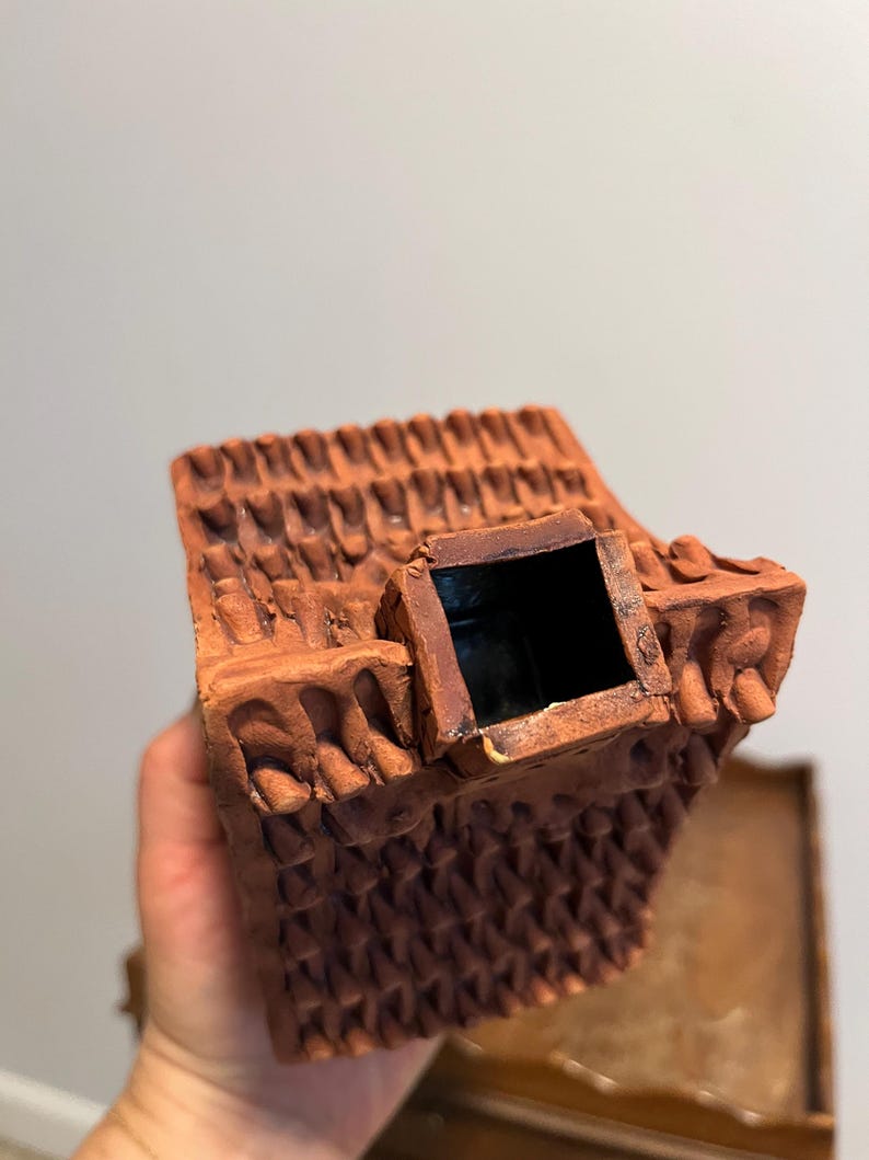 May include: A terracotta-colored ceramic house-shaped container with a textured roof and a square chimney. The container has a dark interior and is held in a person's hand. The house has decorative details.