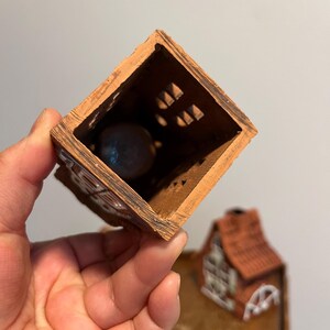 May include: A small, open-topped, brown ceramic house with a red tile roof. The house has decorative cutouts and a dark sphere inside. The house is held in a hand.