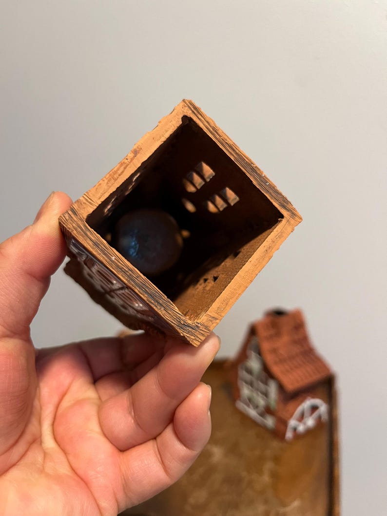 May include: A small, open-topped, brown ceramic house with a red tile roof. The house has decorative cutouts and a dark sphere inside. The house is held in a hand.