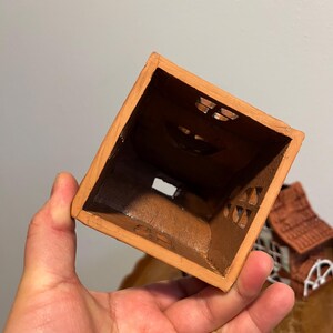 May include: A small, square, terracotta-colored ceramic house with cut-out windows. The interior is dark brown. The house has a rustic, handmade appearance, suggesting it could be a decorative item or a small planter.