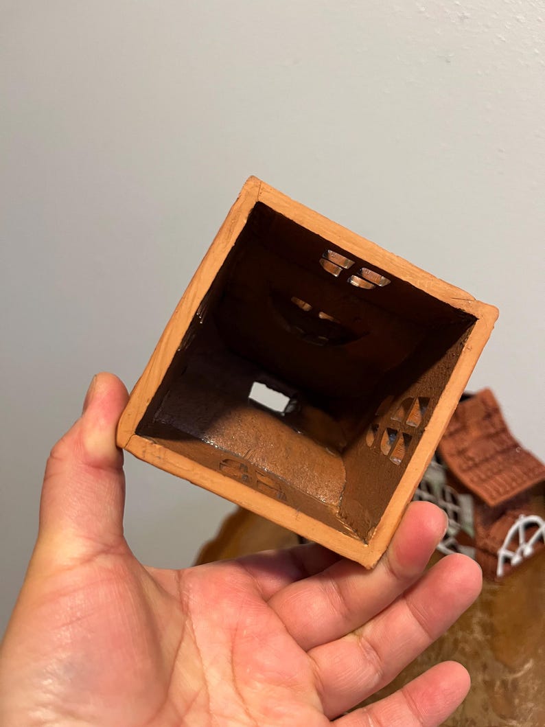 May include: A small, square, terracotta-colored ceramic house with cut-out windows. The interior is dark brown. The house has a rustic, handmade appearance, suggesting it could be a decorative item or a small planter.