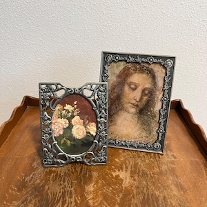 May include: Two silver-toned metal picture frames. The smaller frame is oval-shaped with an ornate floral border containing a print of pink roses. The larger rectangular frame features a detailed relief border and holds a portrait of a person.