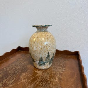 May include: A speckled ceramic vase with a forest scene. The vase has a beige background with black speckles and a green rim. The forest scene features green pine trees. The vase is sitting on a wooden table.