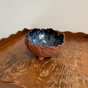 May include: A handmade ceramic bowl with a textured, reddish-brown exterior and a dark blue interior. The bowl has an organic, irregular shape and sits on three small feet. The interior has a glossy finish with gold accents.