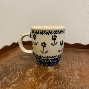 May include: A cream-colored ceramic mug featuring a hand-painted pattern of small dark blue flowers with green stems and leaves. The rim and base are decorated with a dark blue geometric border, and the handle is also adorned with floral motifs.