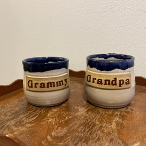 May include: Two ceramic mugs with a light beige base and a dark blue glazed rim. Each mug features a rectangular clay plaque applied to the front with the text "Grammy" on the smaller mug and "Grandpa" on the larger mug in brown lettering.