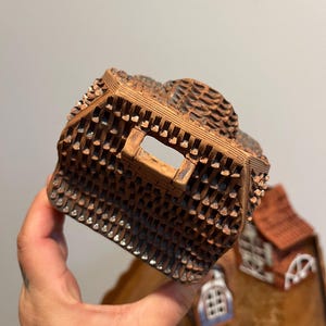 May include: A detailed, brown ceramic miniature house with a textured roof and small, square openings. The house has a rustic appearance, with a small window and a red roof visible in the background. The miniature is held in a hand.