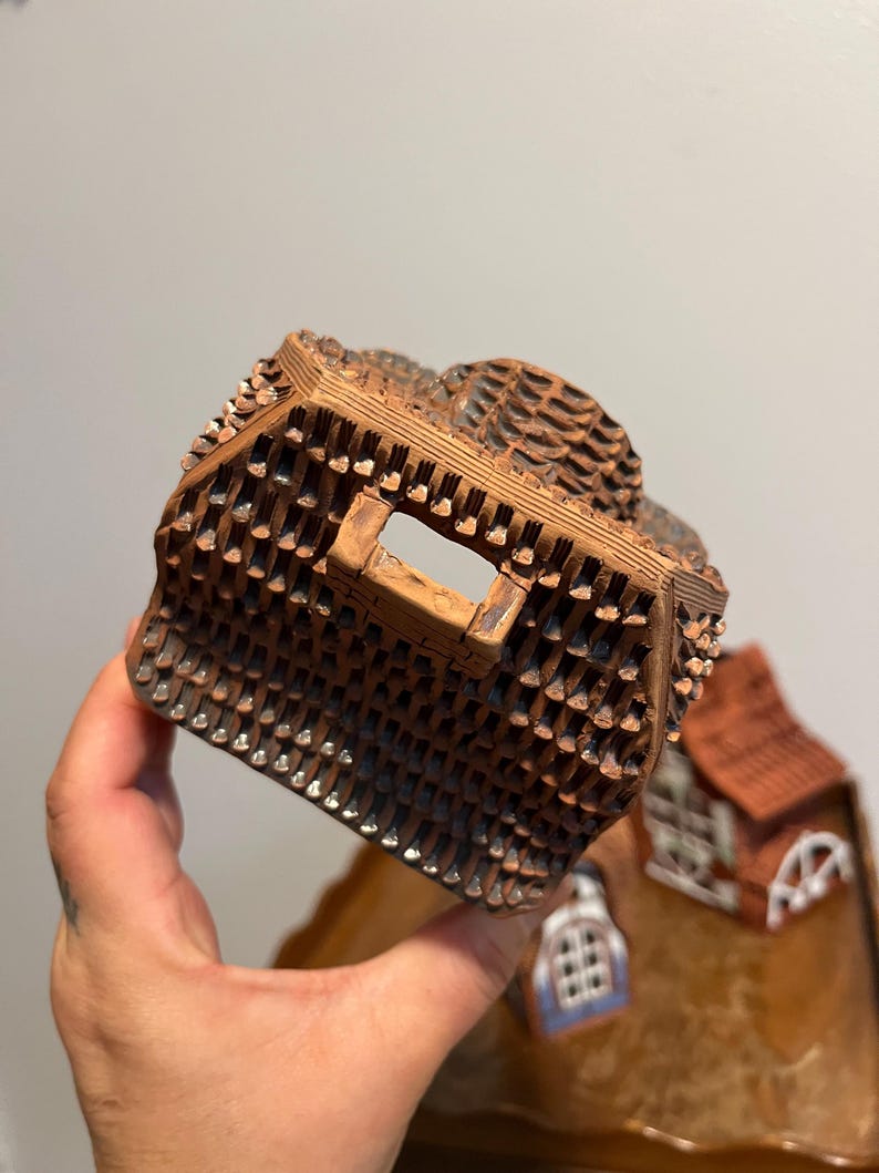 May include: A detailed, brown ceramic miniature house with a textured roof and small, square openings. The house has a rustic appearance, with a small window and a red roof visible in the background. The miniature is held in a hand.