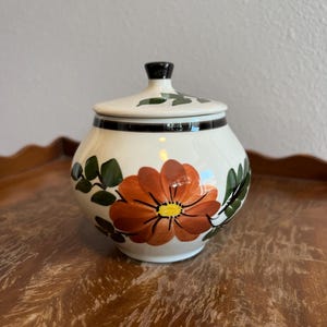 May include: A white ceramic jar with a lid, featuring a large orange flower with a yellow center and green leaves. The jar has a black rim and a black knob on the lid. The floral design is hand-painted.