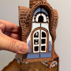 May include: A ceramic house figurine with a brown roof and chimney, and a white and blue window. The roof has a textured, scalloped design. The window has a white frame and a blue border.