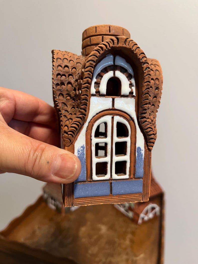 May include: A ceramic house figurine with a brown roof and chimney, and a white and blue window. The roof has a textured, scalloped design. The window has a white frame and a blue border.