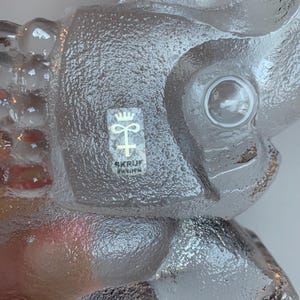 May include: A clear glass elephant figurine with a textured surface. The figurine has a small hole in the top of its head and a sticker on its side that reads "SKRUF SWEDEN".