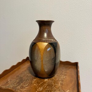 May include: A decorative ceramic vase with a brown, black, and gold color scheme. The vase has a rounded body and a narrow neck. The design features vertical gold and black stripes. The vase is sitting on a wooden table.