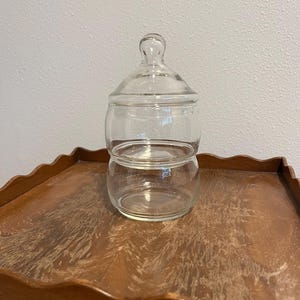 May include: A clear, two-tiered glass jar with a lid. The jar has a rounded shape with a small handle on top of the lid. The jar is sitting on a wooden tray with a scalloped edge. The background is a plain, light-colored wall.