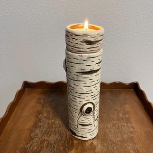 May include: A ceramic candle holder designed to resemble a birch tree trunk. The off-white holder has dark brown markings and a circular design. A lit candle sits inside the top, casting a warm glow. The candle holder is on a wooden table.