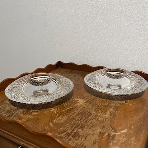 May include: Two clear glass candle holders with a textured, oval design. Each holder has a central well for a candle and sits on a wooden surface. The holders are displayed on a wooden table with a scalloped edge.