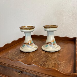 May include: Two ceramic candle holders with a cream base, brown rim, and colorful floral accents. The candle holders are on a wooden table.