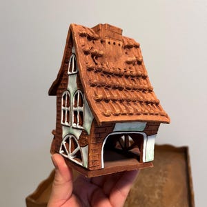 May include: A ceramic house figurine with a brown brick exterior, a red-brown tiled roof, and white-framed windows. The house has an open archway and a chimney. The figurine is held in a hand.