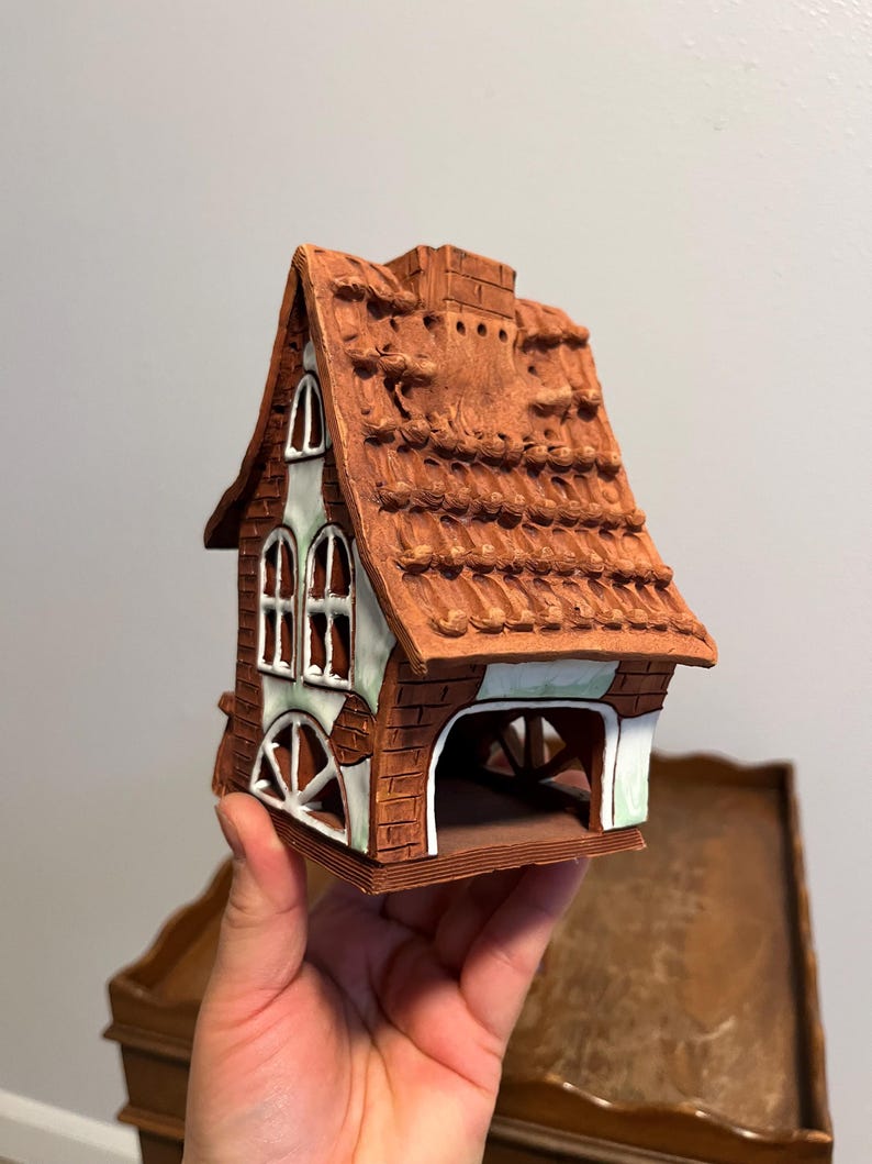 May include: A ceramic house figurine with a brown brick exterior, a red-brown tiled roof, and white-framed windows. The house has an open archway and a chimney. The figurine is held in a hand.