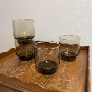May include: Four clear, brown-tinted drinking glasses. Three glasses are arranged on a wooden surface, with one stacked on top of another. The glasses have a simple, cylindrical shape with a thick base.