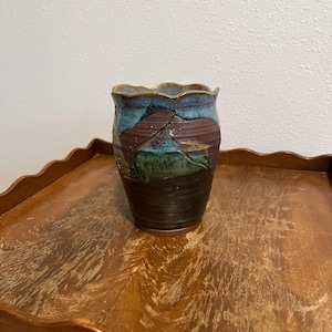 May include: A handmade ceramic vase with a unique, textured design. The vase features a dark brown base, transitioning to shades of green, teal, and brown with a wavy, blue rim. The vase is approximately 5 inches tall.