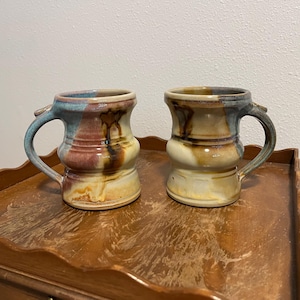 May include: Two ceramic mugs with a flared rim and a rounded midsection. The mugs feature a reactive glaze with blended tones of tan, brown, and blue, showing vertical drip patterns and a smooth, glossy finish with a curved handle on each side.