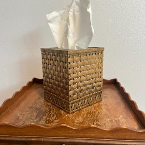 May include: A square tissue box cover featuring a metallic bronze finish with a woven basket-weave texture. The top and bottom edges are accented with a decorative embossed border. A single white tissue extends from the top opening.