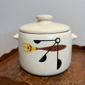 May include: Cream-colored ceramic kitchen canister with lid. The canister features a speckled finish and a graphic of a fork, spoon, and measuring spoons in yellow, brown, and black. The canister has two small handles.
