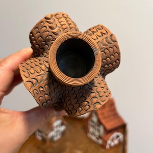 May include: A detailed view of a brown ceramic decorative item. The item has a central circular opening and four textured, petal-like extensions. The surface is intricately patterned with a repeating design. The item is held by a hand.