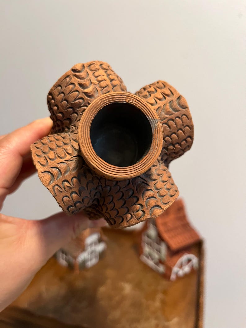 May include: A detailed view of a brown ceramic decorative item. The item has a central circular opening and four textured, petal-like extensions. The surface is intricately patterned with a repeating design. The item is held by a hand.