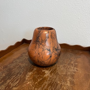 May include: A small, pear-shaped terracotta vase featuring a smooth, polished surface adorned with intricate, dark black, web-like crackle patterns that wrap around the exterior of the vessel, creating a distinct contrast against the warm orange clay.
