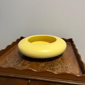 May include: A bright yellow ceramic bowl with a smooth, matte finish and a wide, rounded, donut-like shape. The bowl features a low profile with a circular opening in the center, creating a thick, curved rim that slopes gently toward the interior base.