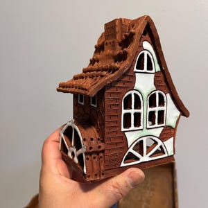 May include: A brown ceramic house with a water wheel, chimney, and arched windows. The roof is textured, and the windows have white and green accents. The house is held in a hand.