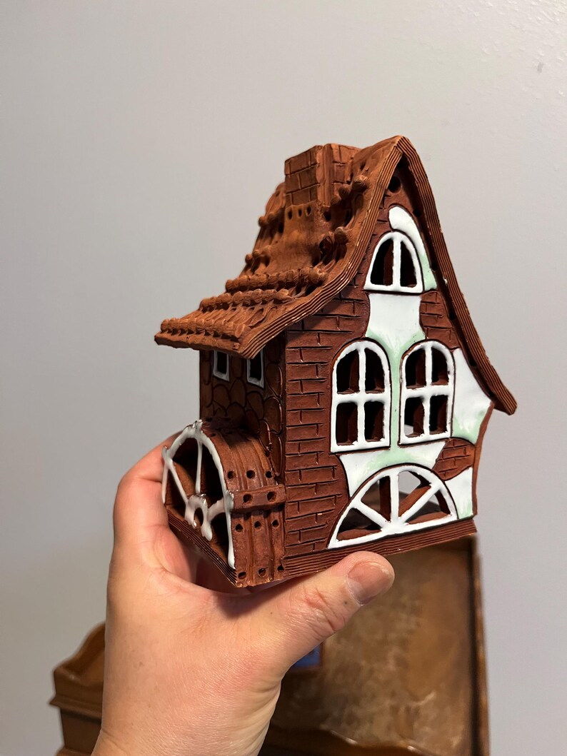 May include: A brown ceramic house with a water wheel, chimney, and arched windows. The roof is textured, and the windows have white and green accents. The house is held in a hand.