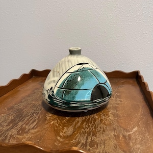 May include: A ceramic vase with a rounded body and a small opening. The vase features a hand-painted design of an igloo in shades of teal, black, and white. The vase sits on a wooden tray.