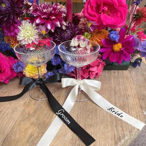 May include: Two champagne flutes with black and white ribbons tied around the stems. The ribbons are labelled "Groom" and "Bride".