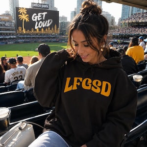 May include: A dark brown hooded sweatshirt featuring the text LFGSD printed in bold yellow letters across the chest. The hoodie includes a front kangaroo pocket and a drawstring hood, providing a casual look suitable for a stadium setting.