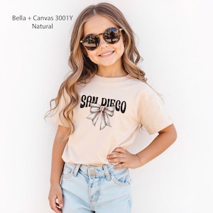 May include: A natural-colored t-shirt with the text "SAN DIEGO" in black, with a baseball-themed bow graphic. The shirt is worn by a person wearing sunglasses and blue jeans. The shirt is made of Bella + Canvas 3001Y Natural material.