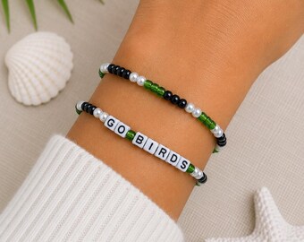 May include: Two beaded bracelets featuring a repeating pattern of small black, white, and green beads. One bracelet includes white square letter beads that spell out the phrase GO BIRDS in black text, while the other consists of simple beads.