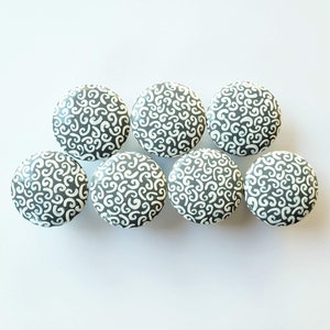 May include: Seven round cabinet knobs with a gray base and a white, swirling pattern. The knobs are evenly spaced against a white background. These decorative knobs can be used for furniture or other home decor projects.