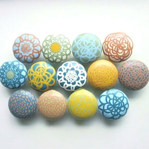 Może przedstawiać: Kolekcja dwunastu dekoracyjnych ceramicznych gałek w różnych kolorach, w tym niebieskim, żółtym, pomarańczowym i zielonym. Każda gałka ma unikalny kwiatowy lub geometryczny wzór, idealny do mebli.