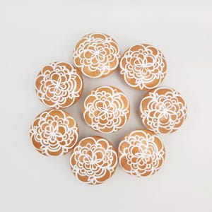 May include: Overhead shot of eight round, light brown cabinet knobs arranged in a circle. Each knob features a white, floral design. The knobs are set against a plain, white background, creating a clean and elegant aesthetic.