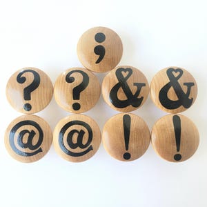 May include: A collection of wooden cabinet knobs with black painted punctuation marks: question marks, ampersands, exclamation points, at signs, and a semicolon. The knobs are arranged on a white surface.
